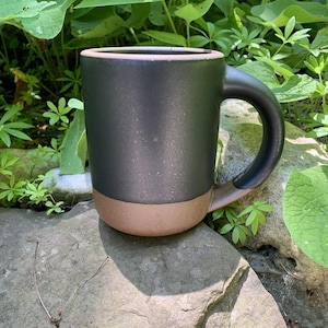 May include: A cylindrical ceramic mug featuring a two-tone design with a matte black upper section and a contrasting unglazed terracotta-colored base. The mug has a sturdy, curved handle that matches the black finish of the upper body.