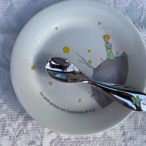 Puede incluir: Un plato de cerámica blanco con una representación del Principito de pie en un asteroide, rodeado de estrellas. El plato tiene el texto "Le petit prince sur l'astéroïde B 612." Una cuchara de plata con una estrella verde en el mango está apoyada en el plato.
