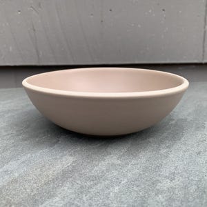 May include: A light pink ceramic bowl with a rounded shape. The bowl has a smooth, matte finish and is set against a gray background. This versatile bowl is suitable for serving various dishes.
