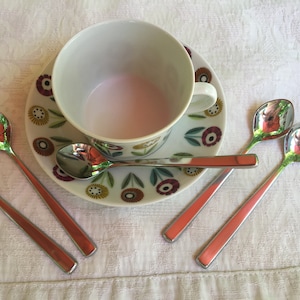 May include: Four silver teaspoons with rounded bowls and square handles, arranged around a white teacup and saucer with a floral pattern. The teacup is filled with a light pink liquid.