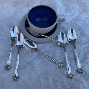 May include: Six silver spoons with ornate handles arranged on a white tablecloth with a blue ceramic teacup and saucer in the background.
