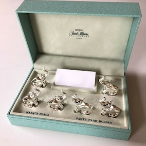 May include: A set of six silver-toned elephant-shaped place card holders presented in a light blue textured box. The interior features the text Ercuis Saint Hilaire Collection Paris, Marque Place, and Party Card Holder with a blank white card.