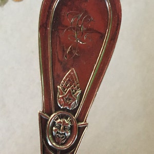 Pode incluir: Uma colher de prata com um cabo ornamentado com uma cabeça de carneiro, uma flor estilizada e um rosto em um círculo. A colher tem uma pátina vermelho-marrom.