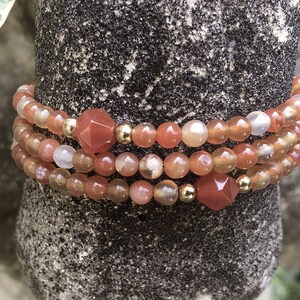 Könnte beinhalten: Ein mehrreihiges Armband aus Karneolperlen in Orange- und Rottönen. Das Armband hat goldene Zwischenstücke und größere Karneolperlen mit einer facettierten Oberfläche.