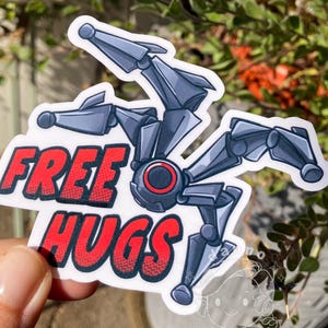 May include: A sticker featuring a gray robot spider with the words "FREE HUGS" in red and white. The robot has a red circular center and multiple articulated legs. The sticker is held against a background of green foliage.