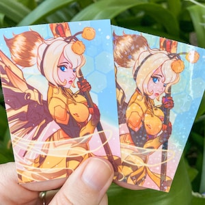 May include: Two holographic stickers featuring a cartoon character with blonde hair, yellow and orange clothing, and a staff. The character is surrounded by a blue and pink background with a honeycomb pattern.
