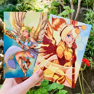 May include: Two illustrated cards featuring female characters. The first card has a light blue background with a woman with blonde hair and wings, holding a staff. The second card has a yellow background with a woman with blonde hair and wings, holding a staff.