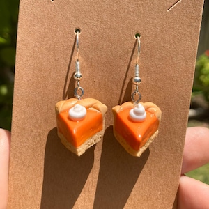 May include: A pair of dangle earrings featuring a miniature pumpkin pie slice with a white dollop of whipped cream. The earrings are made of polymer clay and have silver hooks.