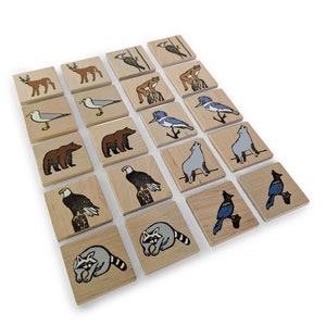May include: A set of twenty square wooden tiles arranged in a grid, each featuring a unique illustration of North American wildlife including bears, eagles, raccoons, deer, seagulls, blue jays, and wolves, rendered in a simple, graphic style.