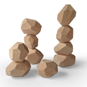 May include: A collection of light-colored wooden blocks shaped like irregular, multi-faceted stones. The blocks are stacked into two vertical towers of varying heights, with one additional block resting on the surface in the foreground.