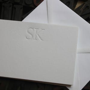 Pode incluir: Um cart&atilde;o branco com um monograma em relevo das letras "SK" em uma fonte simples.