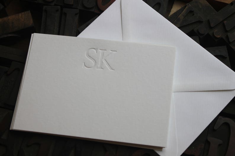 Pode incluir: Um cart&atilde;o branco com um monograma em relevo das letras "SK" em uma fonte simples.