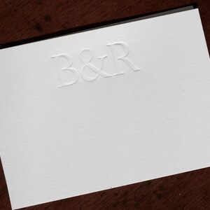 Pode incluir: Um cart&atilde;o branco com as letras B&R em relevo na parte superior central do cart&atilde;o.