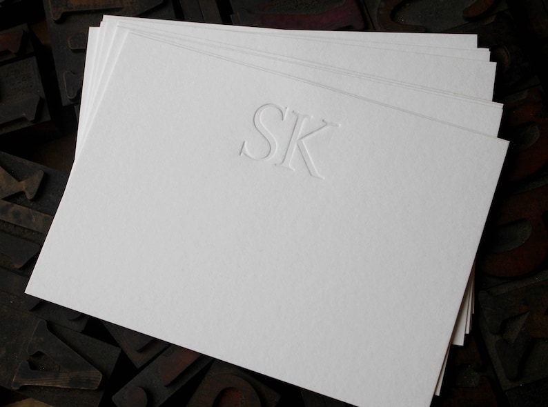 Pode incluir: Um conjunto de cart&otilde;es de papelaria brancos com as iniciais "SK" em relevo no centro.