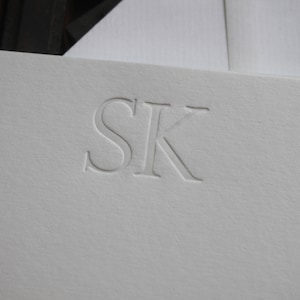 Pode incluir: Papel&atilde;o branco com as iniciais "SK" em relevo em uma fonte simples e elegante.