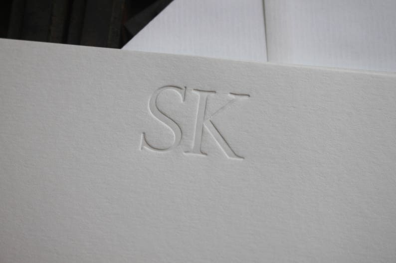 Pode incluir: Papel&atilde;o branco com as iniciais "SK" em relevo em uma fonte simples e elegante.