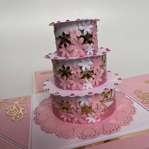 May include: A three-tiered pink and white cake with gold accents and floral embellishments. The cake is decorated with pink and white flowers and gold glitter. The cake is sitting on a white doily with a pink background.