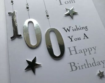 Può includere: Un biglietto bianco con lettere e decorazioni argentate. Il biglietto dice "To Captain Tom Wishing You A Happy Birthday". Il numero 100 è scritto in grandi lettere argentate.