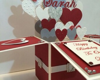 May include: A red and white pop-up card with a heart design. The card is open and the inside says "Happy Birthday to Our Sarah".
