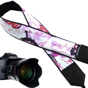 May include: A white camera strap with a pink floral pattern and black text. The strap has black padding on both ends and a black InTePro logo. The strap is designed for a camera.
