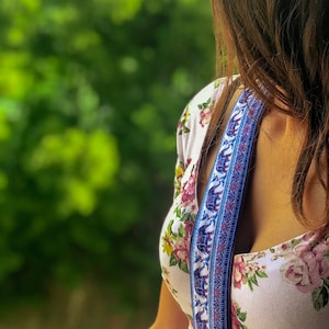 May include: A woman wearing a white floral dress with a blue and white patterned camera strap. The strap features a repeating pattern of elephants.