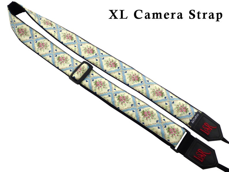 May include: A white and blue floral camera strap with black adjustable straps and the text "LSR" embroidered in red on the strap ends. The strap is labeled "XL Camera Strap".
