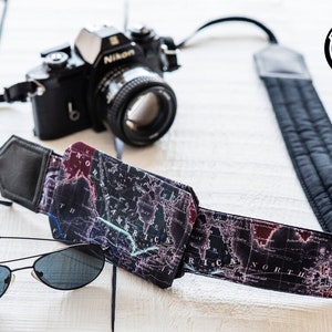 May include: A black camera with a black and white map patterned camera strap. The strap has a black leather section and a black padded section. A pair of black sunglasses are lying on a white wooden surface.