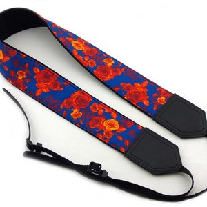May include: A blue camera strap with a floral pattern featuring red and orange roses. The strap has black leather ends with adjustable straps.