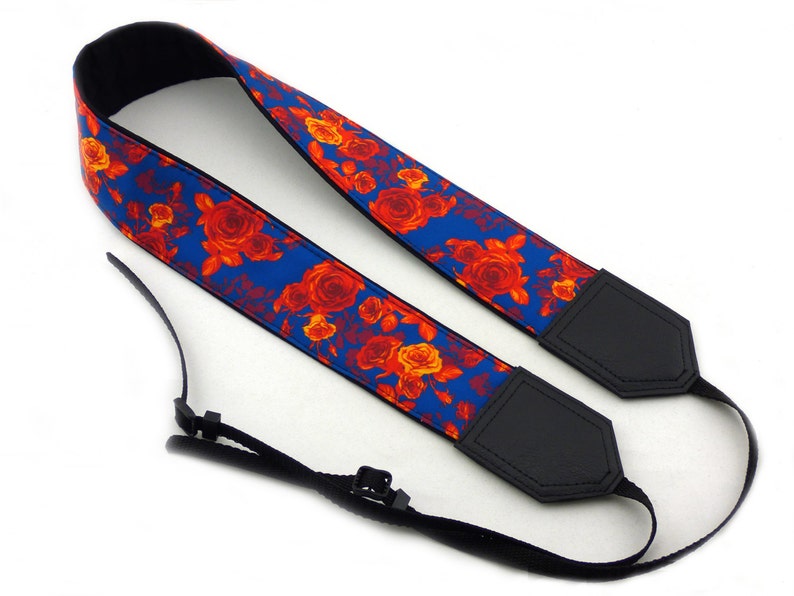 May include: A blue camera strap with a floral pattern featuring red and orange roses. The strap has black leather ends with adjustable straps.