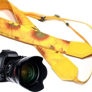Op de afbeelding: Een gele cameraband met een zonnebloemprint. De band heeft twee verstelbare gevulde delen. Een zwarte DSLR-camera met een lens is op de achtergrond.