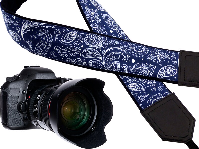 May include: A blue and white paisley camera strap with black leather accents. The strap is adjustable and has a padded shoulder section for comfort.
