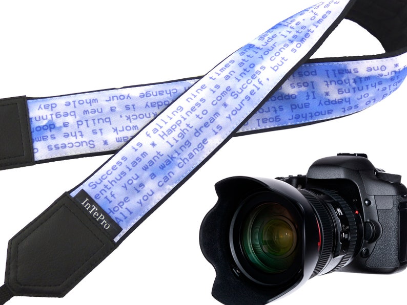 May include: A blue and white camera strap with the text "Success is falling nine times and getting up ten. Happiness is an attitude. If you want light to come into your dream, all you can change is yourself. But sometimes success consists of only one small step, or happy and strong stories. To set another goal." The strap is attached to a black camera with a large lens.