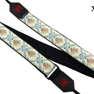 May include: A white and blue floral guitar strap with black padding and a black adjustable buckle. The strap has the letters "LSR" embroidered in red on both ends. The strap is labeled "XL".