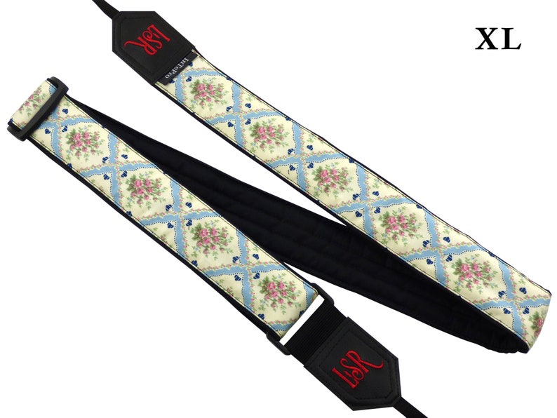 May include: A white and blue floral guitar strap with black padding and a black adjustable buckle. The strap has the letters "LSR" embroidered in red on both ends. The strap is labeled "XL".