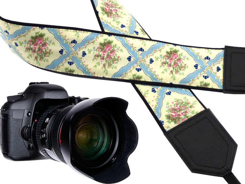 May include: A camera strap with a floral pattern in shades of pink, blue, and yellow. The strap is made of fabric and has a black leather trim. The strap is shown with a black camera.