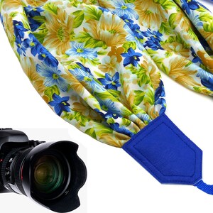 Pode incluir: Uma alça de câmera floral azul e branca com uma almofada de couro azul. A alça é feita de tecido e tem um padrão floral. A alça está presa a uma câmera preta.