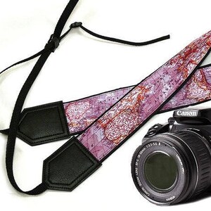 May include: A pink and brown camera strap with a world map design. The strap has black leather accents and is attached to a black camera with a lens.