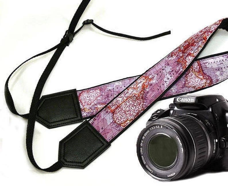 May include: A pink and brown camera strap with a world map design. The strap has black leather accents and is attached to a black camera with a lens.