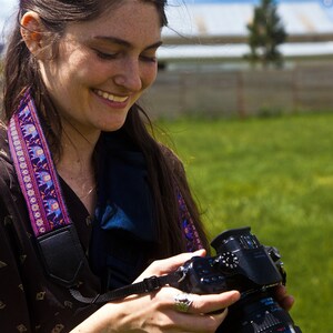 May include: A woman with long brown hair is holding a black camera with a lens. She is wearing a brown and black patterned shirt with a purple and blue patterned camera strap.