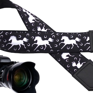 May include: Black and white camera strap with a repeating pattern of white horses and horseshoes. The strap has black leather ends.