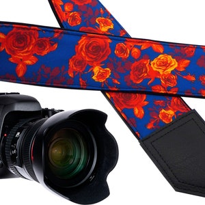May include: A blue and red floral camera strap with black leather accents. The strap is made of fabric and has a floral pattern with red roses on a blue background. The strap has a black leather adjustable strap and a black leather pad.