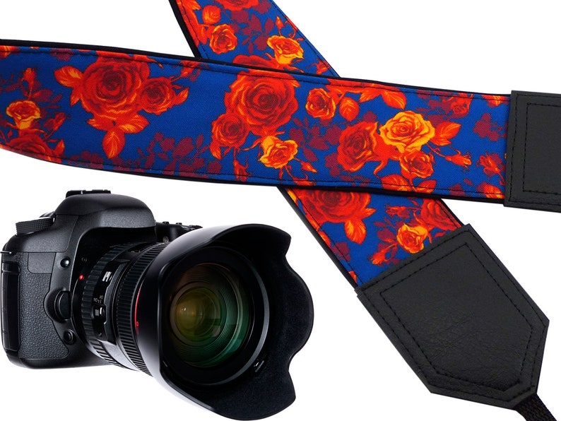 May include: A blue and red floral camera strap with black leather accents. The strap is made of fabric and has a floral pattern with red roses on a blue background. The strap has a black leather adjustable strap and a black leather pad.