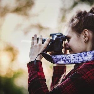 May include: A woman in a red and black plaid shirt is holding a black camera with a map-patterned strap and taking a picture.