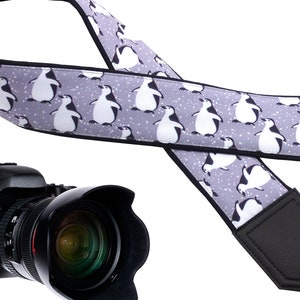 May include: A gray camera strap with a penguin pattern and black leather accents. The strap is designed to be worn over the shoulder.