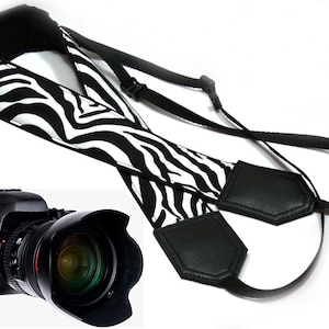 May include: A black and white zebra patterned camera strap with black leather accents. The strap is adjustable and has a quick release buckle.