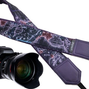 May include: A purple camera strap with a world map design and the text "Intrepid" printed on the strap. The strap is attached to a black camera with a lens.