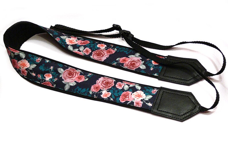 May include: A black camera strap with a floral pattern featuring pink roses on a dark blue background. The strap has black leather ends.