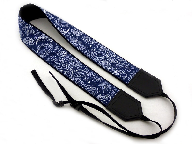 May include: A blue and white paisley camera strap with black adjustable straps and black leather ends.