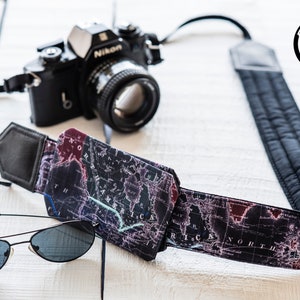 May include: A black camera strap with a map print design. The strap has a black leather pad and a black camera with a silver lens. A pair of black sunglasses are also in the image.