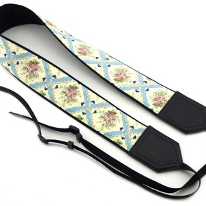 May include: A camera strap with a floral pattern in shades of pink, yellow, and blue. The strap is made of fabric and has black leather accents.